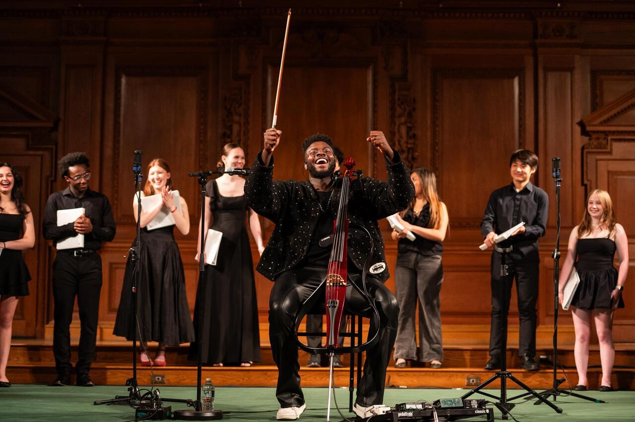  Kevin Olusola ’06 performs on cello at center stage in Cochran Chapel at Phillips Academy, raising a bow while students stand behind holding sheet music.